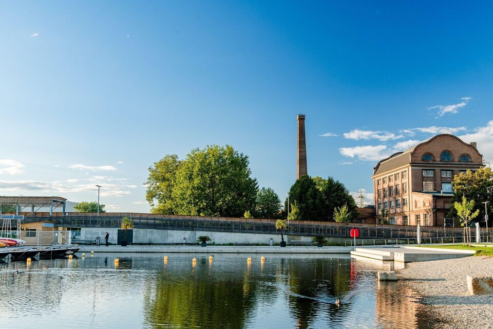 Ein ruhiger Wasserbereich mit einem historischen Gebäude und einem alten Schornstein im Hintergrund. Grüne Bäume und ein klarer Himmel vervollständigen die Szenerie.
