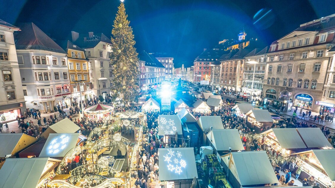 Ein festlicher Weihnachtsmarkt mit vielen Lichtern und Ständen. Im Hintergrund steht ein großer, beleuchteter Weihnachtsbaum. | © Stadt Graz Foto Fischer
