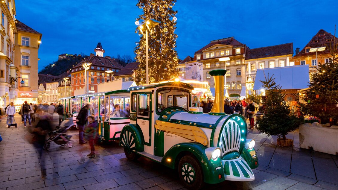 Ein festlich geschmückter Platz mit einem beleuchteten Weihnachtsbaum und einem kleinen Zug. Viele Menschen schlendern durch die weihnachtliche Atmosphäre. | © Graz Tourismus-Harry Schiffer