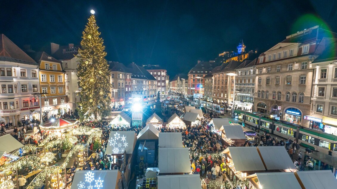Ein festlicher Weihnachtsmarkt bei Nacht mit einem großen, beleuchteten Weihnachtsbaum. Die Stände sind voller Lichter und Menschen, die die Atmosphäre genießen. | © Stadt Graz Foto Fischer