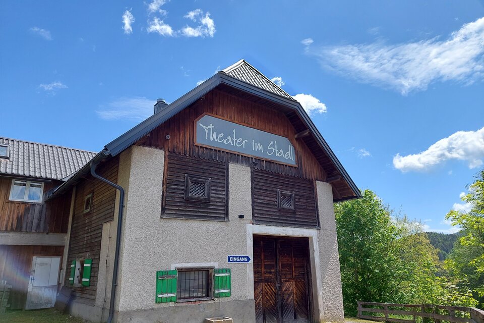 Ein charmantes Gebäude mit einem Schild, das „Theater im Stadel“ zeigt. Der Himmel ist blau mit einigen Wolken und es gibt viel Grün um das Gebäude herum. | © Brigitte Digruber