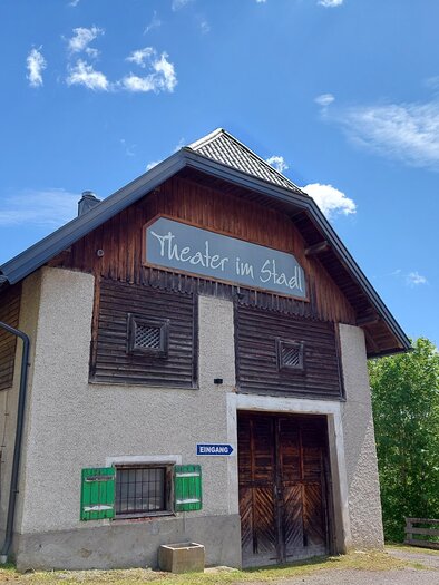 Ein charmantes Gebäude mit einem Schild, das „Theater im Stadel“ zeigt. Der Himmel ist blau mit einigen Wolken und es gibt viel Grün um das Gebäude herum. | © Brigitte Digruber