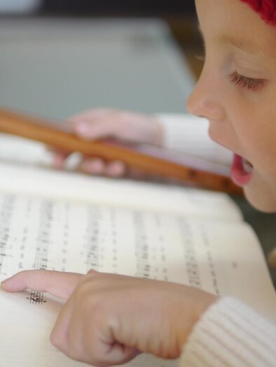 Ein kleines Mädchen spielt Flöte und liest aus einem Musikbuch. Sie trägt eine rote Mütze und zeigt auf die Noten. | © Steirisches Volksliedwerk