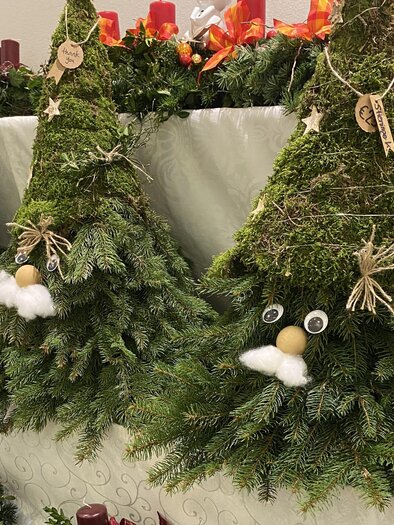 Drei dekorative Weihnachtsbäume aus Moos und Tannenzweigen stehen auf einem Tisch. Jeder Baum hat ein lustiges Gesicht mit einem weißen Bart und verschiedenen Anhängern. | © Eva-Maria Paier