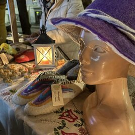 A shop window with a classic mannequin wearing a purple felt hat. In the background, handcrafted shoes and other goods are displayed on a table. | © Oststeiermark Tourismus