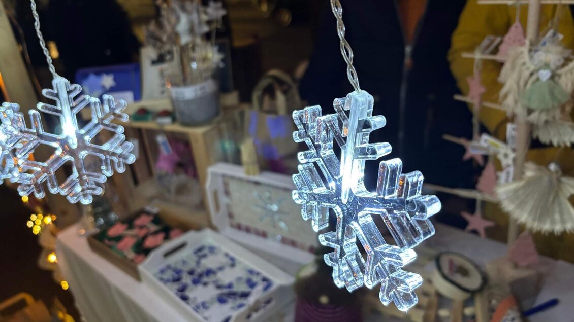 Eine festliche Lichterdekoration mit glitzernden Schneeflocken hängt über einem Verkaufsstand. Der Hintergrund zeigt verschiedene handgefertigte Produkte in warmem Licht. | © Oststeiermark Tourismus