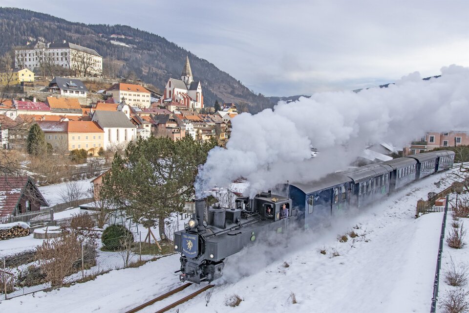 Eine dampfbetriebene Lokomotive fährt durch eine verschneite Landschaft. Im Hintergrund sind malerische Häuser und eine Kirche zu sehen. | © Tv Murau