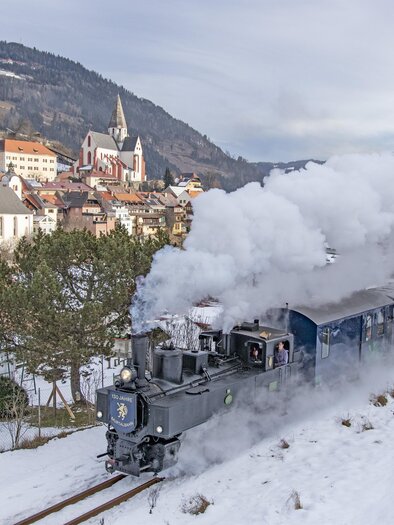 Eine dampfbetriebene Lokomotive fährt durch eine verschneite Landschaft. Im Hintergrund sind malerische Häuser und eine Kirche zu sehen. | © Tv Murau