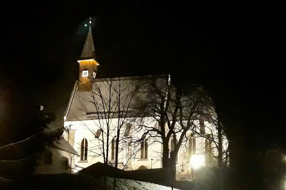 Eine beleuchtete Kirche bei Nacht, umgeben von Bäumen. Der Himmel ist dunkel, und die Atmosphäre ist ruhig. | © Gitti Fladl