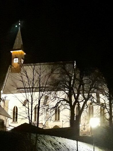 Eine beleuchtete Kirche bei Nacht, umgeben von Bäumen. Der Himmel ist dunkel, und die Atmosphäre ist ruhig. | © Gitti Fladl
