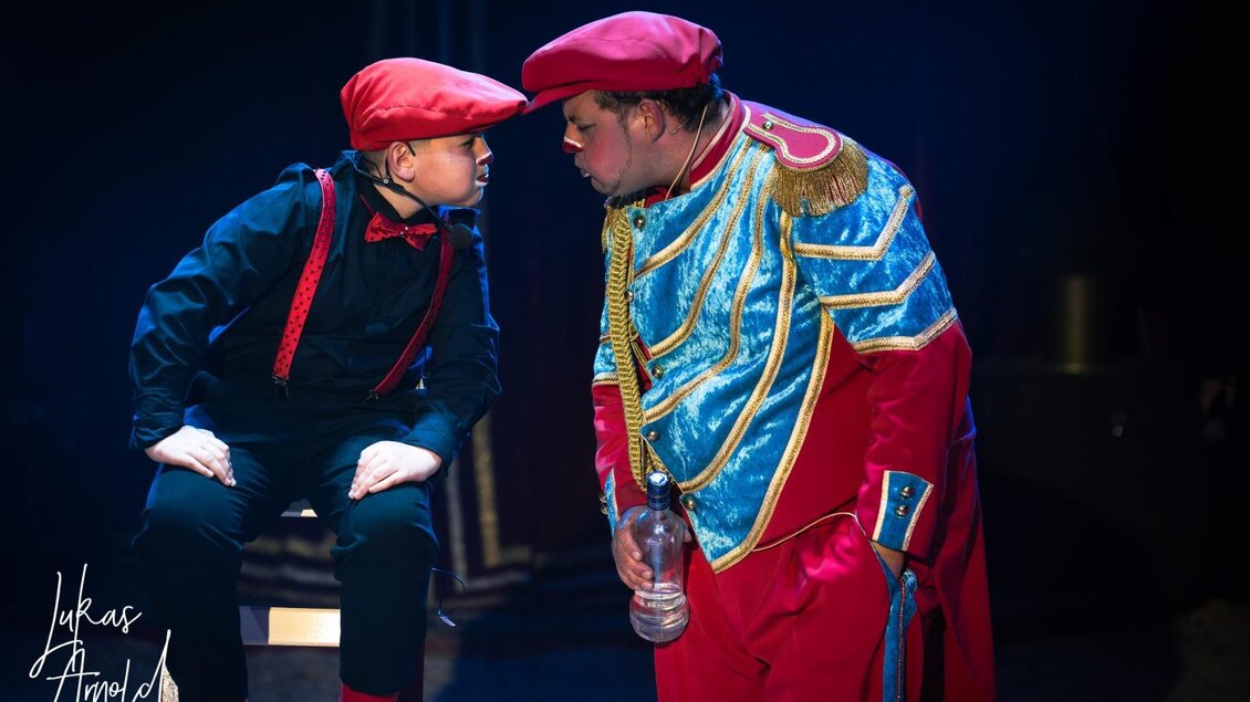 Zwei Clowns mit roten Mützen stehen sich in der Manege gegenüber. Einer trägt eine blau-rote Uniform und hält eine Flasche. | © Circus Frankello-Lukas Arnold
