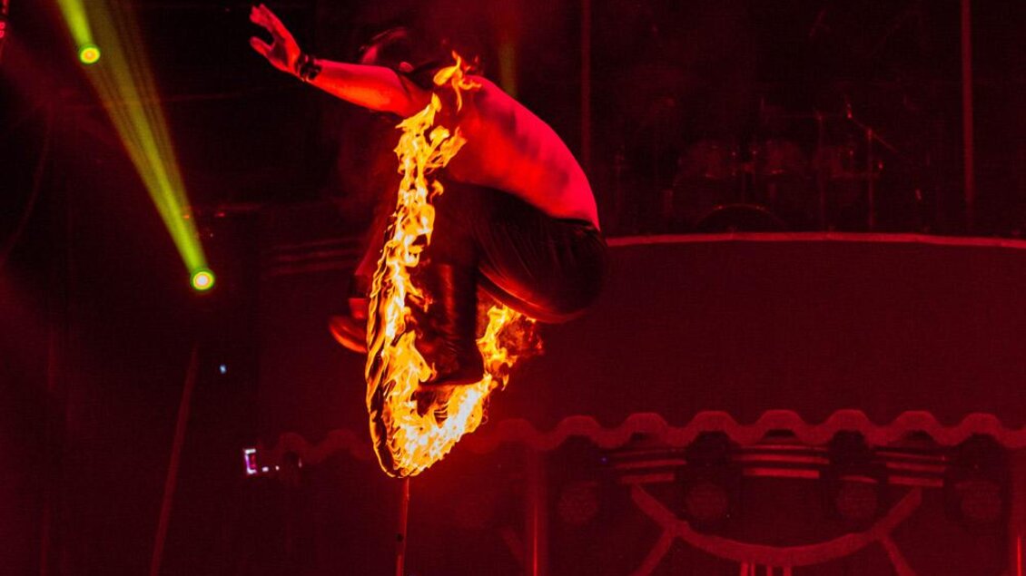 Ein Akrobat springt mit angezogenen Beinen durch einen brennenden Reifen in der rot beleuchteten Zirkusmanege. | © Circus Frankello-Lukas Arnold