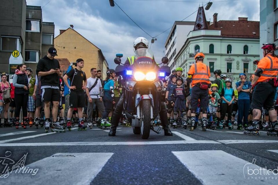 Eine Gruppe von Menschen, die auf Inline-Skates stehen, versammelt sich an einer Straße. Ein Polizeimotorrad mit eingeschaltetem Blaulicht steht vorne und sorgt für Sicherheit während der Veranstaltung.