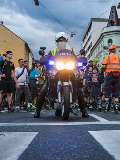 Eine Gruppe von Menschen, die auf Inline-Skates stehen, versammelt sich an einer Straße. Ein Polizeimotorrad mit eingeschaltetem Blaulicht steht vorne und sorgt für Sicherheit während der Veranstaltung.
