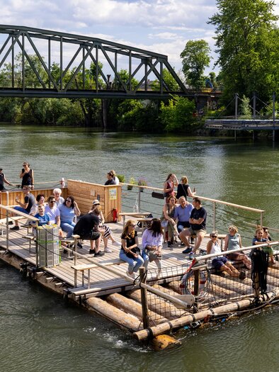 Eine Gruppe von Menschen sitzt auf einem Floss im Wasser. Im Hintergrund ist eine Brücke und Bäume zu sehen. | © die flösserei-Harry Schiffer
