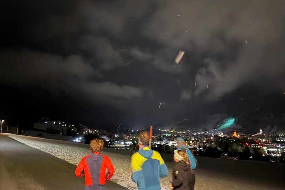 Drei Läufer joggen nachts auf einer Straße, umgeben von Schnee und beleuchteten Bergen. Der Himmel ist dunkel mit Wolken. | © we.move