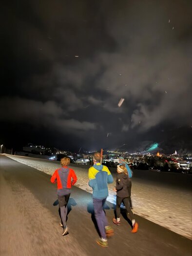 Drei Läufer joggen nachts auf einer Straße, umgeben von Schnee und beleuchteten Bergen. Der Himmel ist dunkel mit Wolken. | © we.move