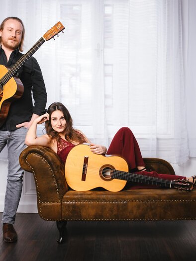 Ein Mann und eine Frau stehen und liegen auf einer Couch und halten Gitarren. Sie blicken entspannt in die Kamera, umgeben von hellen Vorhängen. | © @Lukas Bezila