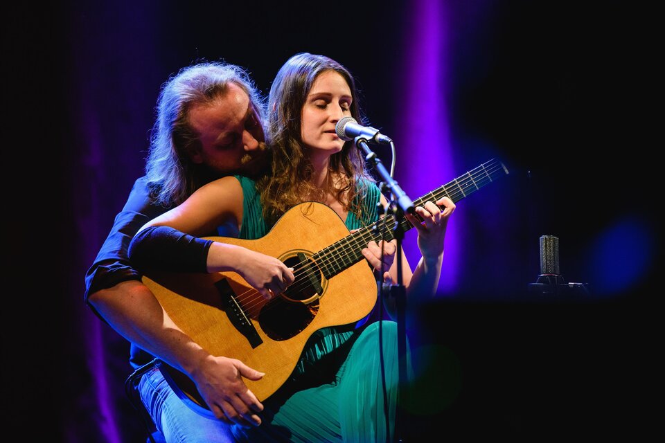 Ein Musikerpaar führt ein Lied auf der Bühne vor. Die Frau spielt Gitarre und singt, während der Mann sie von hinten umarmt. | © Lukas Bezila
