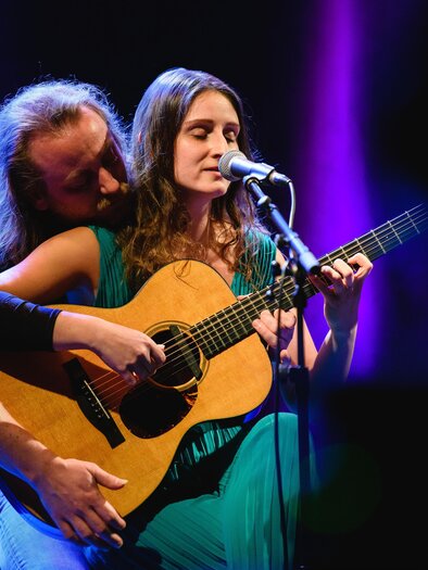 Ein Musikerpaar führt ein Lied auf der Bühne vor. Die Frau spielt Gitarre und singt, während der Mann sie von hinten umarmt. | © Lukas Bezila