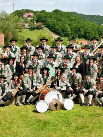 Trachtenmusikkapelle Gossendorf in traditioneller Uniform vor südoststeirischer Hügellandschaft – gelebte Blasmusik und starke Gemeinschaft in der Erlebnisregion Thermen- & Vulkanland.