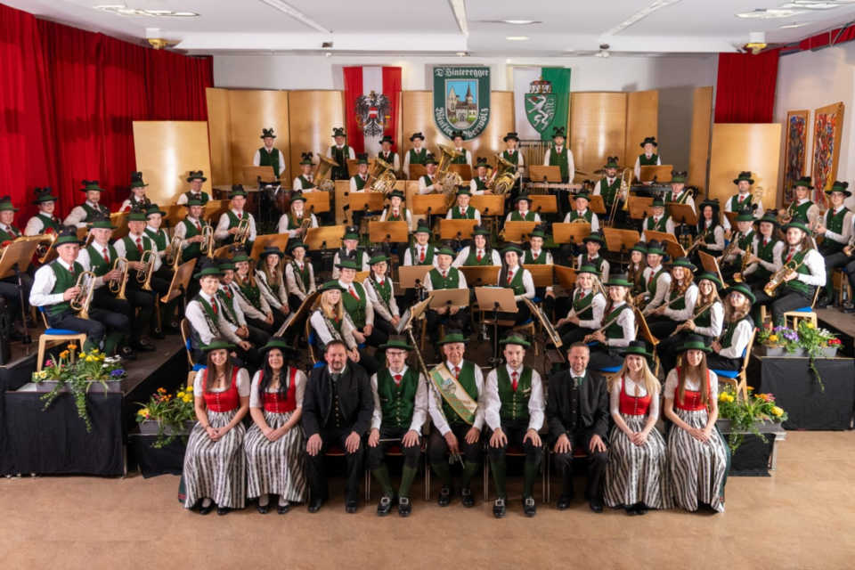 Eine große Musikkapelle steht auf der Bühne, umgeben von bunten Fahnen und Dekorationen. Die Musiker tragen traditionelle Kleidung und haben eine festliche Ausstrahlung. | © TV Murau