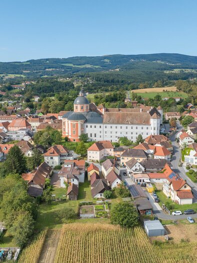 Eine malerische Stadt mit roten Dächern und einer beeindruckenden Kirche im Mittelpunkt. Im Hintergrund sind grüne Hügel und ein blauer Himmel zu sehen. | © Helmut Schweighofer