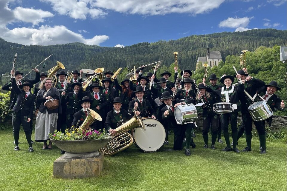 Eine große Gruppe von Musikern steht im Freien und lächelt. Sie tragen traditionelle Trachten und halten verschiedene Musikinstrumente.