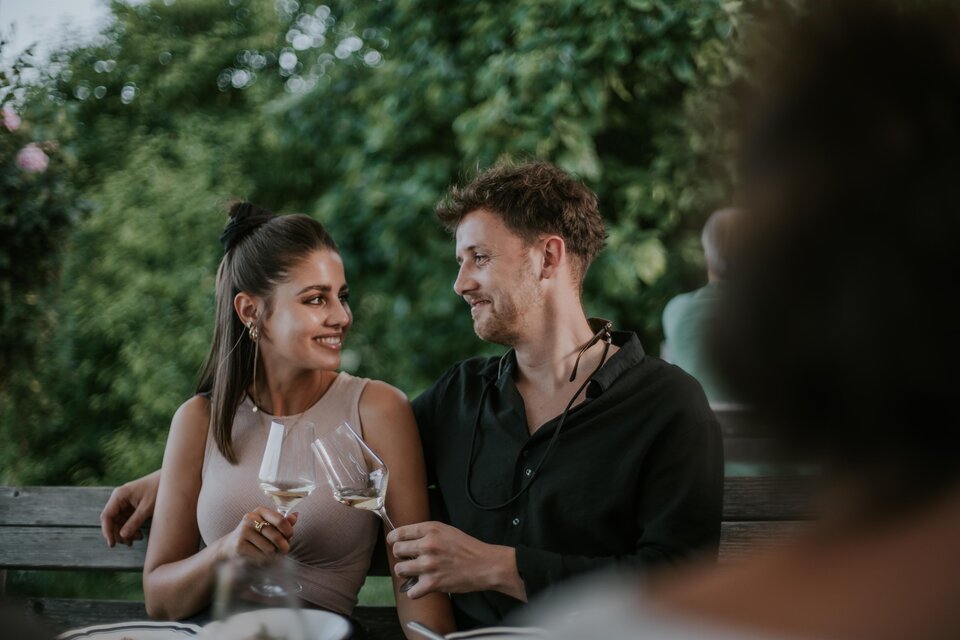 Ein Paar sitzt an einem Tisch im Freien und schaut sich verliebt an. Beide halten Weingläser und genießen den Moment. | © TVB Südsteiermark – Lukas Elsneg