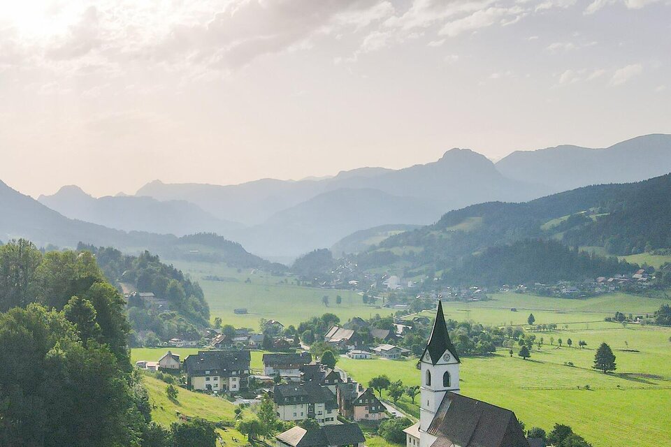 Eine malerische Landschaft mit einem kleinen Dorf und einer Kirche. Im Hintergrund sind sanfte Hügel und ein klarer Himmel zu sehen. | © TV Gesäuse