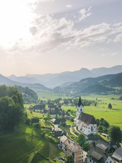 Eine malerische Landschaft mit einem kleinen Dorf und einer Kirche. Im Hintergrund sind sanfte Hügel und ein klarer Himmel zu sehen. | © TV Gesäuse