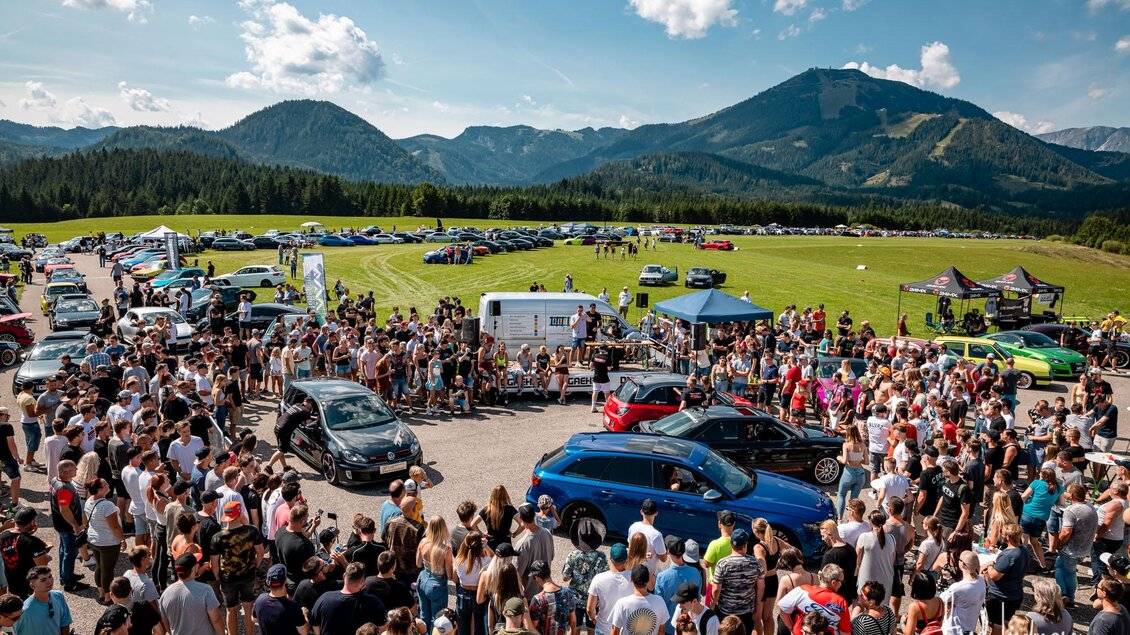 Ein großes Auto-Event mit einer großen Menschenmenge. Im Hintergrund sind grüne Berge und blue Himmel zu sehen. | © www.mariazell.blog