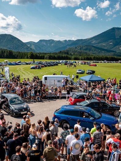 Ein großes Auto-Event mit einer großen Menschenmenge. Im Hintergrund sind grüne Berge und blue Himmel zu sehen. | © www.mariazell.blog