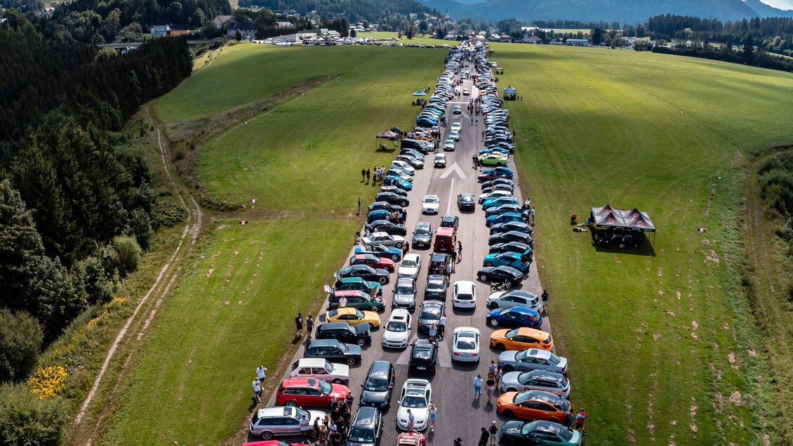 Eine lange Reihe von Autos auf einer Straße in einer ländlichen Umgebung. Im Hintergrund sind grüne Wiesen und Berge zu sehen. | © www.mariazell.blog