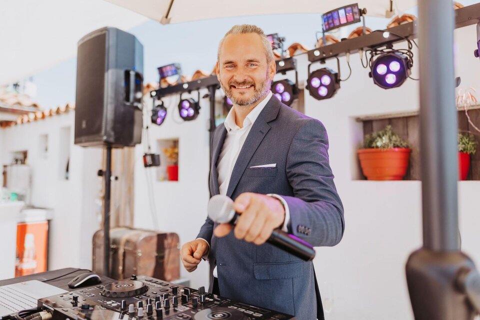 Ein DJ in einem eleganten Anzug steht vor einem Mischpult und lächelt. Im Hintergrund sind Lichter und eine festliche Atmosphäre zu sehen. | © Daniel Pack DJ