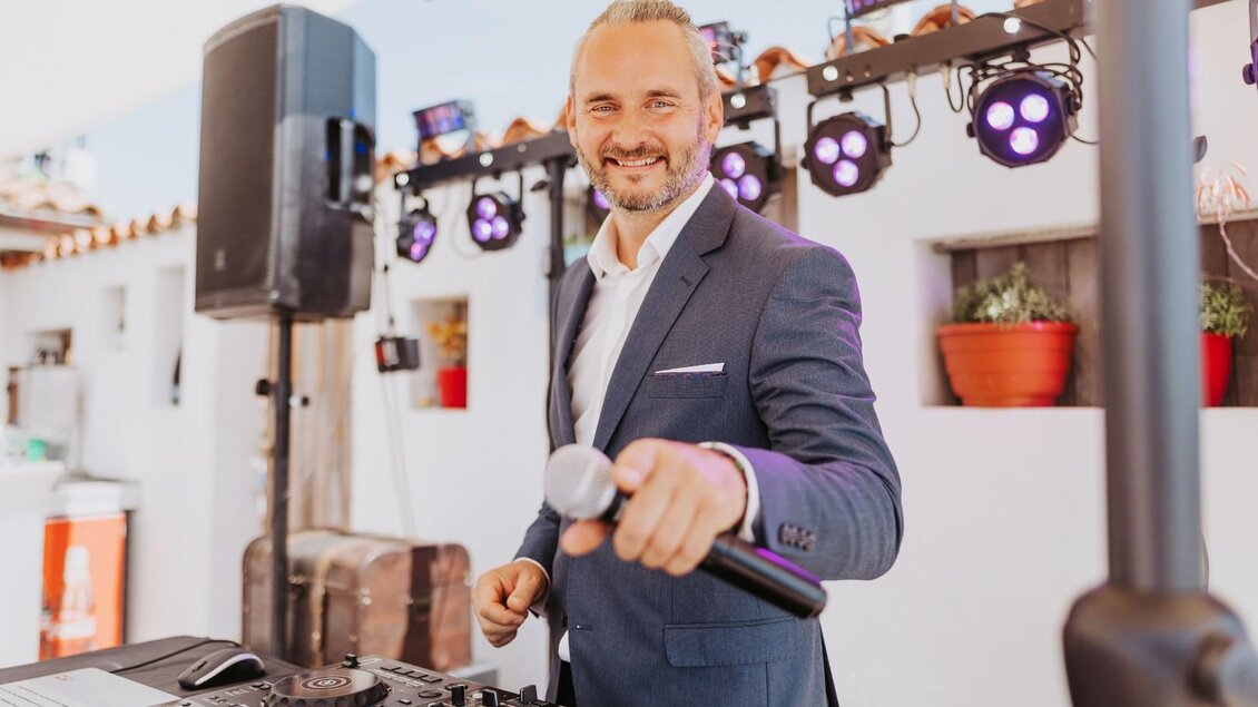 Ein DJ in einem eleganten Anzug steht vor einem Mischpult und lächelt. Im Hintergrund sind Lichter und eine festliche Atmosphäre zu sehen. | © Daniel Pack DJ