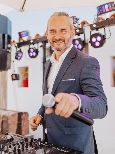 Ein DJ in einem eleganten Anzug steht vor einem Mischpult und lächelt. Im Hintergrund sind Lichter und eine festliche Atmosphäre zu sehen. | © Daniel Pack DJ