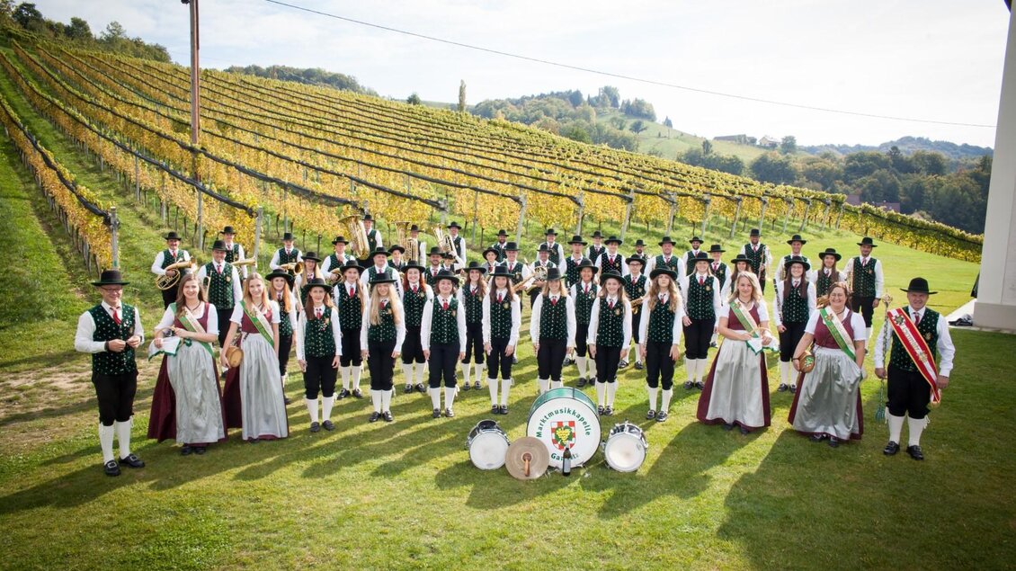 Eine Gruppe von festlich gekleideten Menschen steht in einem Weinberg. Im Hintergrund sind die Weinreben und ein klarer Himmel zu sehen. | © Marktmusikkapelle Gamlitz