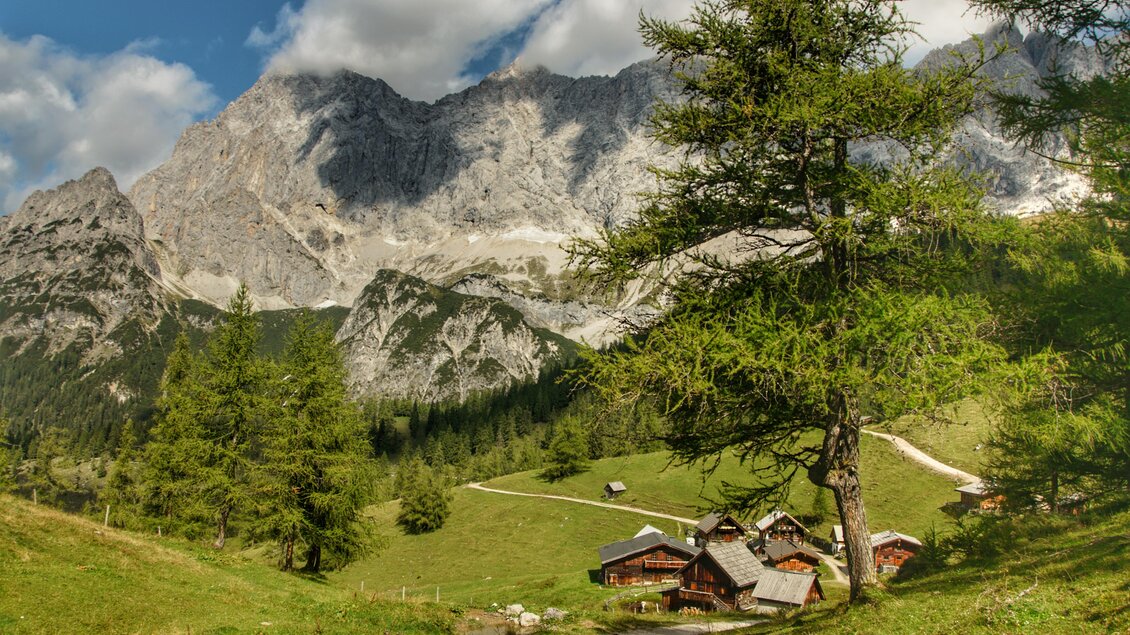 Eine malerische alpine Landschaft mit hohen Bergen und grünen Wiesen. Traditionelle Holzhäuser sind am Fuß der Berge zu sehen. | © Marlene Eggmayr