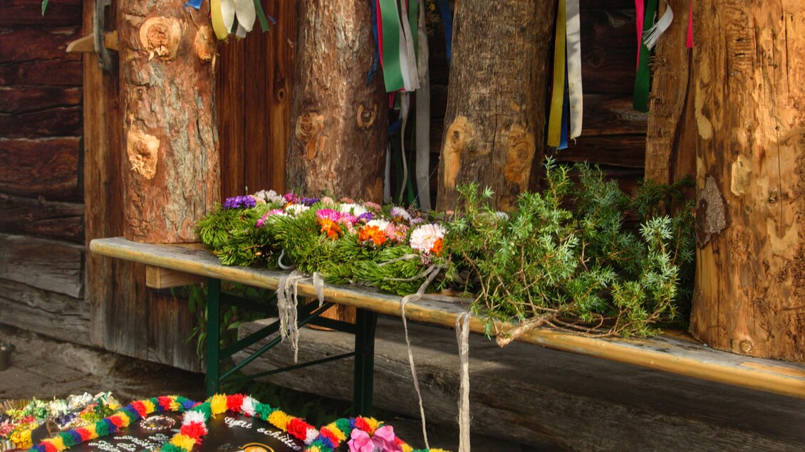 Eine Holzbank mit bunten, handgefertigten Dekorationen liegt vor einer Holzwand. Darüber hängen Blumenkränze und Bänder in verschiedenen Farben. | © Marlene Eggmayr