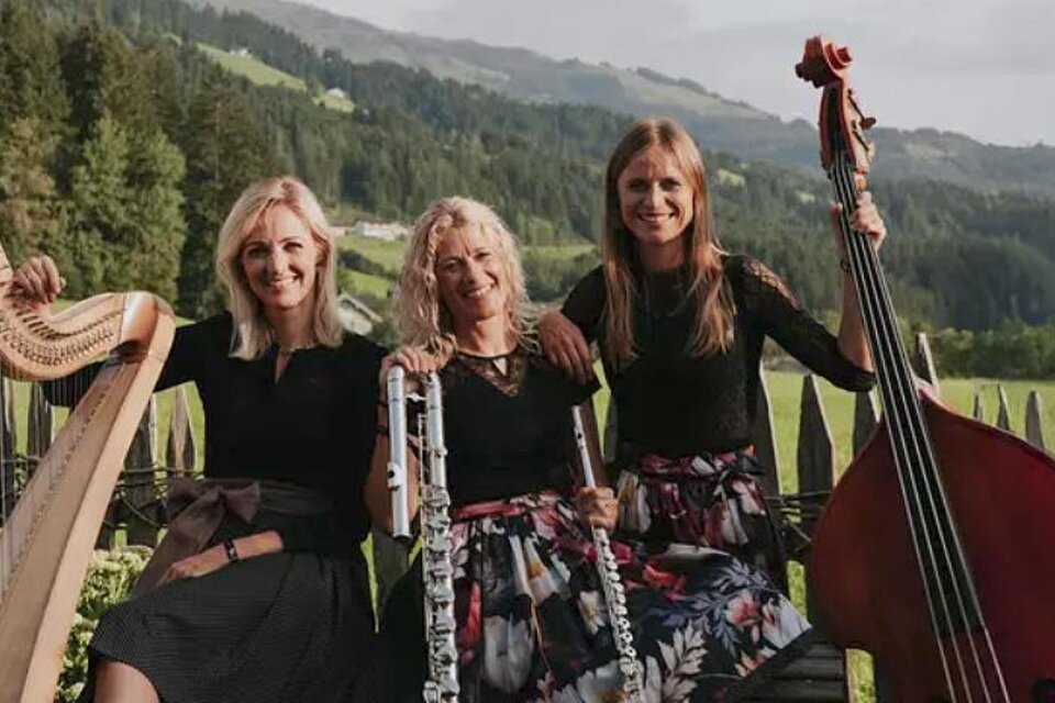 Drei Musikerinnen stehen in einer malerischen Landschaft. Sie spielen verschiedene Instrumente, darunter eine Harfe und ein Kontrabass.