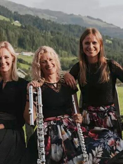 Drei Musikerinnen stehen in einer malerischen Landschaft. Sie spielen verschiedene Instrumente, darunter eine Harfe und ein Kontrabass.
