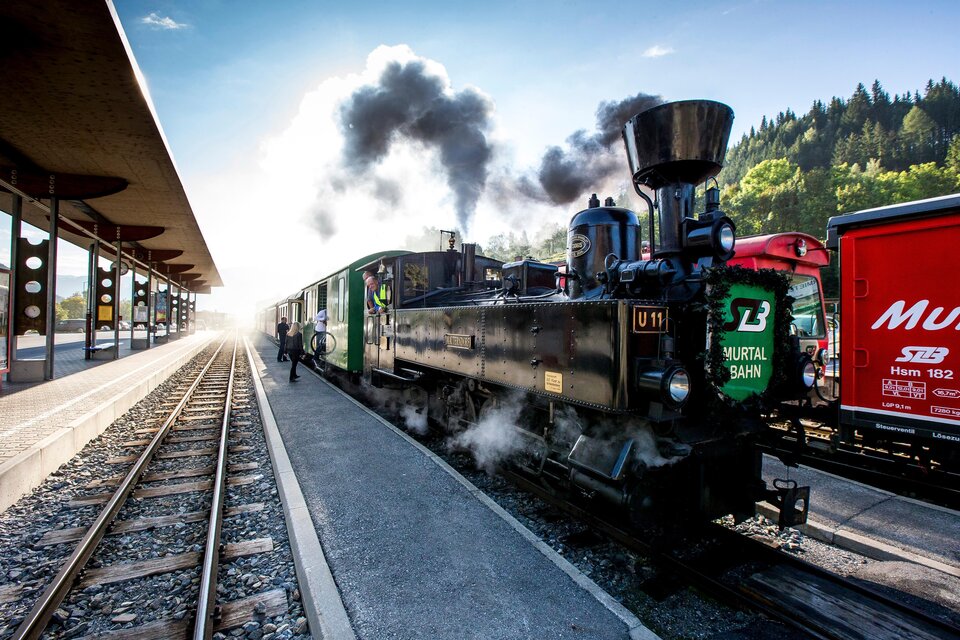 Eine Dampflok steht auf dem Bahnhof, Rauch steigt aus dem Schornstein auf. Im Hintergrund sind weitere Waggons und eine bewaldete Landschaft zu sehen.