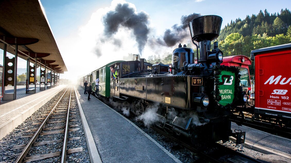 Eine Dampflok steht auf dem Bahnhof, Rauch steigt aus dem Schornstein auf. Im Hintergrund sind weitere Waggons und eine bewaldete Landschaft zu sehen.