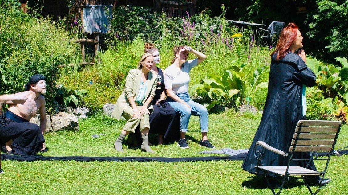 <p>Das Ensemble der Bluatschwitz Black Box führt im Garten voller Pflanzen eine Vorstellung auf. Einige Personen posieren, während eine andere Person in einem schwarzen Mante geradel spricht.</p> | © Bluatschwitzblackbox, bluatschwitzblackbox.eu/