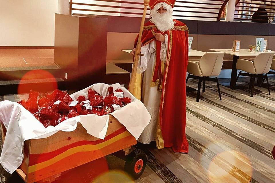 Ein gekleideter Mann als Nikolaus steht neben einem Wagen voller Geschenke. Die Szene spielt sich in einem modernen Innenraum ab. | © H2O Kindertherme