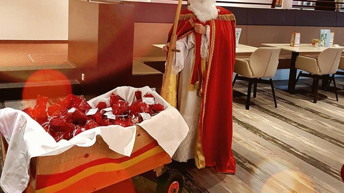 Ein gekleideter Mann als Nikolaus steht neben einem Wagen voller Geschenke. Die Szene spielt sich in einem modernen Innenraum ab. | © H2O Kindertherme