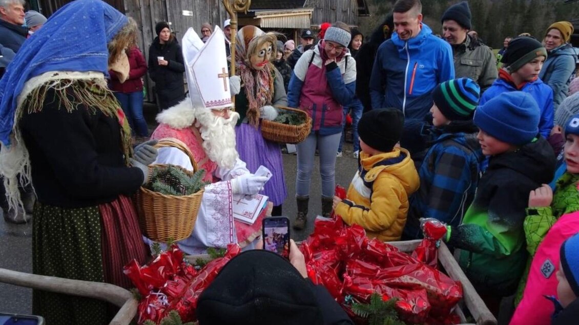 Eine festliche Szene mit einer Person im Kostüm des Nikolaus, die von Kindern umgeben ist. Im Hintergrund gibt es Menschen und Geschenke in roten Taschen. | © Johanna Rastl