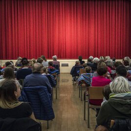 Ein voll besetzter Saal mit Menschen, die auf die Bühne schauen. Der rote Vorhang ist gespannt und wartet auf die Vorstellung. | © Tv Murau