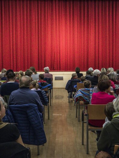 Ein voll besetzter Saal mit Menschen, die auf die Bühne schauen. Der rote Vorhang ist gespannt und wartet auf die Vorstellung. | © Tv Murau